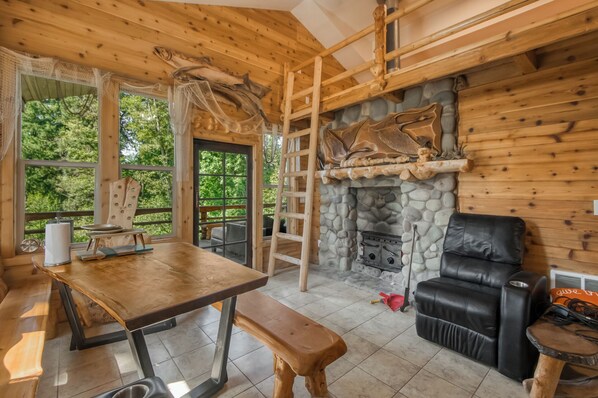 Interior rústico de una cabaña con chimenea de piedra, mobiliario de madera y vistas al bosque.