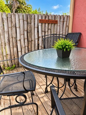 A round glass table with metal chairs, a potted plant, and a bamboo fence with a sign.