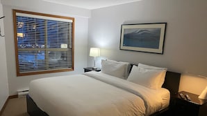 A hotel room with a large bed, a nightstand, a lamp, a framed picture on the wall, and a window with blinds.