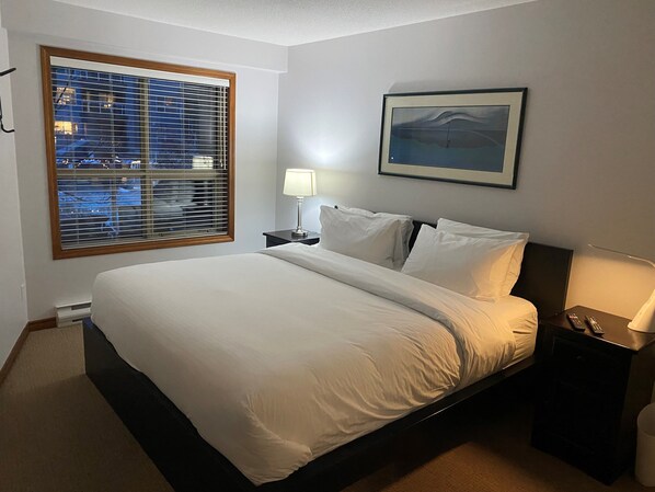 A hotel room with a large bed, a nightstand, a lamp, a framed picture on the wall, and a window with blinds.