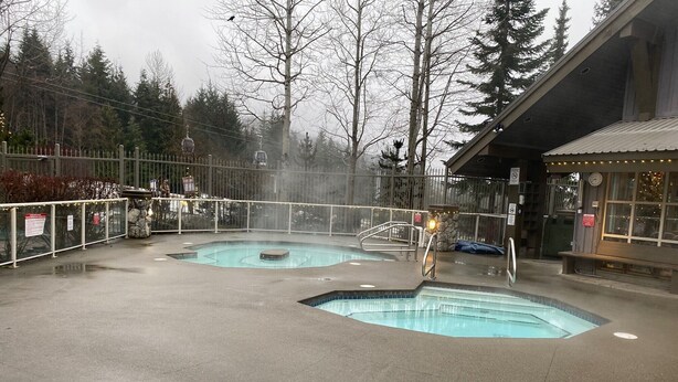A pool area with two hot tubs, a stone wall, and a wooden building with lights.