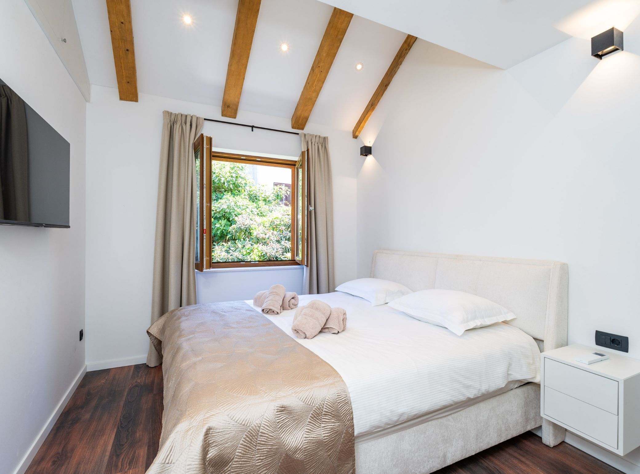A bedroom with a bed, a nightstand, a TV mounted on the wall, and a window with curtains.