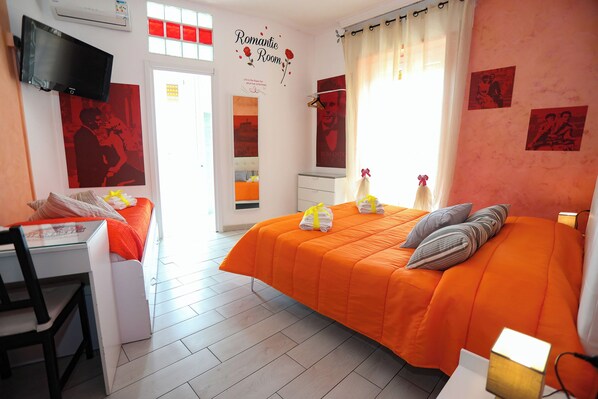 A bedroom with an orange bedspread, a desk with a chair, a flat-screen TV, and a wall with a 'Romantic Room' sign.