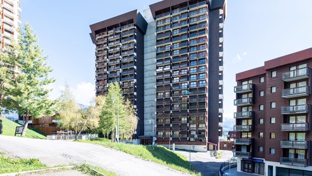 Ein modernes Mehrfamilienhaus mit mehreren Balkonen, einer gepflasterten Fläche davor und blauem Himmel.