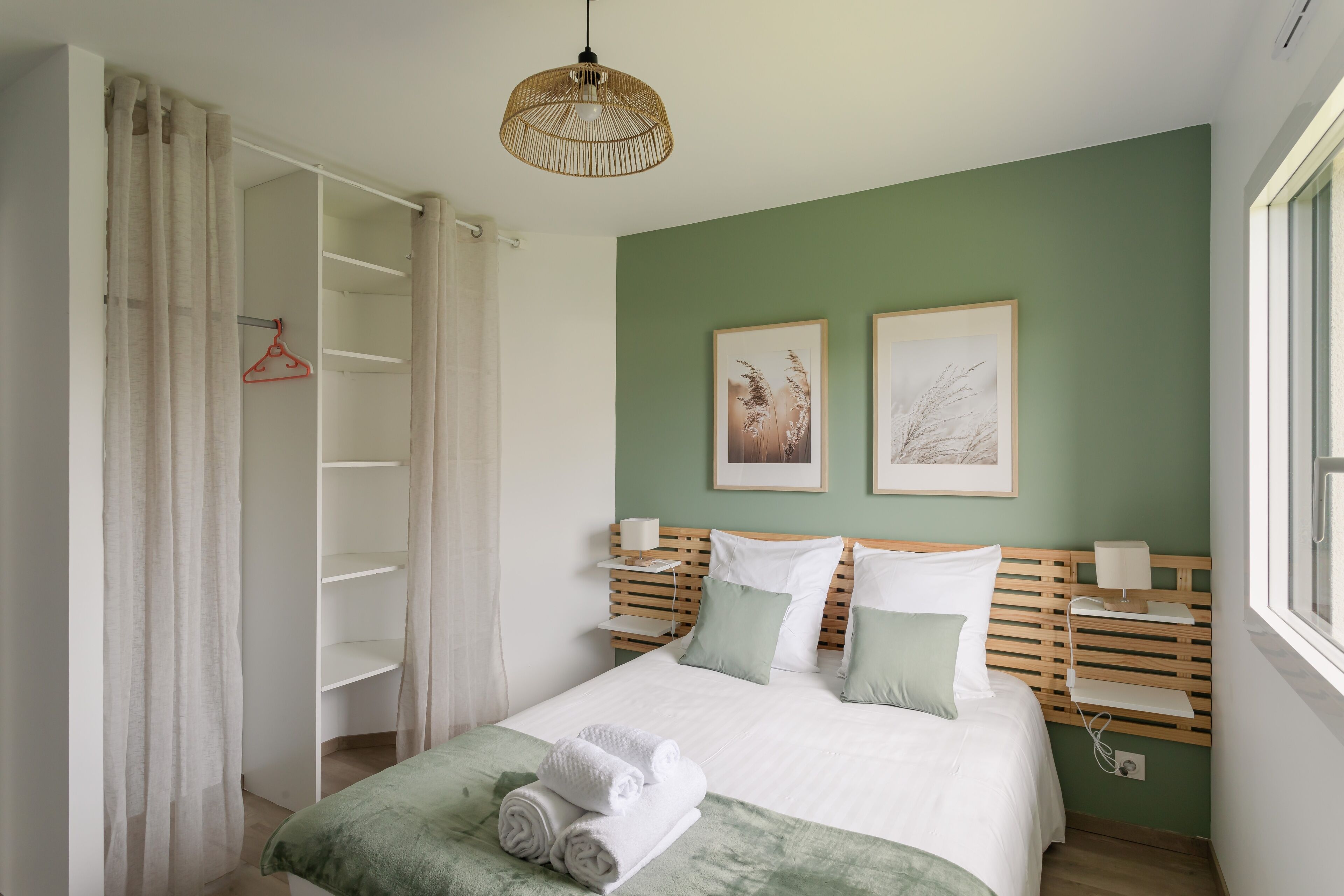 A neatly made bed with a green bedspread, two pillows, and rolled towels. There is a bedside table with a lamp, a wooden headboard, and two framed pictures on the wall. A closet with a hanging rack is visible to the left.