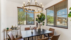 Uma sala de jantar com uma mesa redonda, seis cadeiras, um candelabro e grandes janelas com persianas.