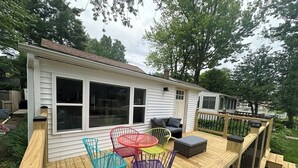 En terrasse med farverige stole og et lille bord, en trægelænder og et hvidt hus med vinduer.