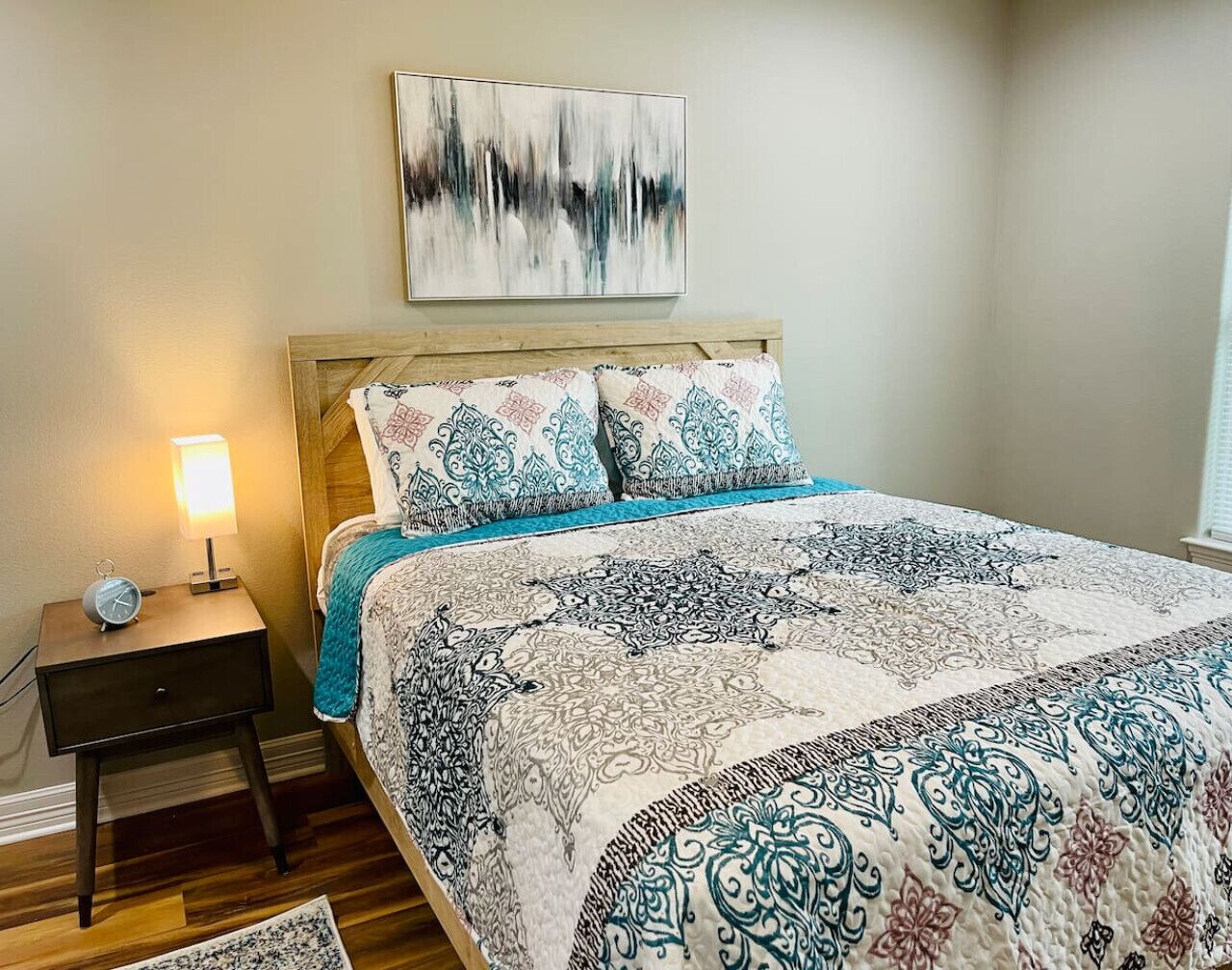 A bedroom with a wooden headboard, a patterned bedspread, a nightstand with a lamp, and a wall-mounted abstract painting.