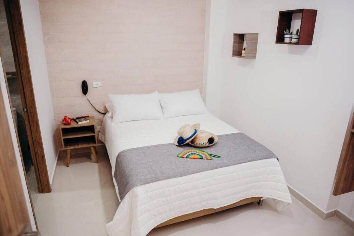 Un letto rifatto con un copriletto bianco e una coperta grigia, un piccolo comodino in legno con una lampada, e una mensola a muro con oggetti decorativi.
