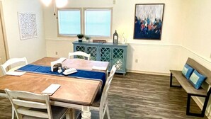 Un coin repas avec une table en bois, des chaises blanches, un chemin de table bleu, un buffet bleu et un banc avec des coussins bleus.