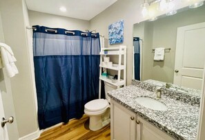 Un baño con inodoro blanco, cortina de ducha azul, lavamanos de granito y estante blanco con artículos de tocador.