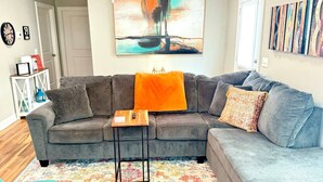 Un salon avec un canapé d’angle gris, une petite table d’appoint en bois et un tapis coloré.