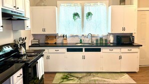 A modern kitchen with white cabinets, a black stove, a microwave, and a window with curtains. - Bright & Airy 2BR 1BA Home Near The Riverfront (Port Neches)