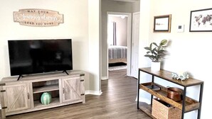 A living room with a TV on a wooden stand, a console table with baskets, and a wall sign.