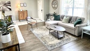 A living room with a sofa, coffee table, and chairs. There is a wooden cabinet, a plant, and a window with curtains.
