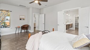 A spacious bedroom with a large bed, a ceiling fan, and a view of the kitchen through an open door. - Perfect house for vacation or work w/Patio, Yard & Parking! 5 min from Kalahari! (Round Rock)