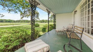 A covered porch with a view of a green landscape, featuring a bench and a chair. - Farmhouse at Magnolia Valley (Fort Valley)