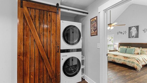 A laundry room with two stacked front-loading washers and dryers, a wooden barn-style door, and a bedroom with a bed and a ceiling fan. - Farmhouse at Magnolia Valley (Fort Valley)