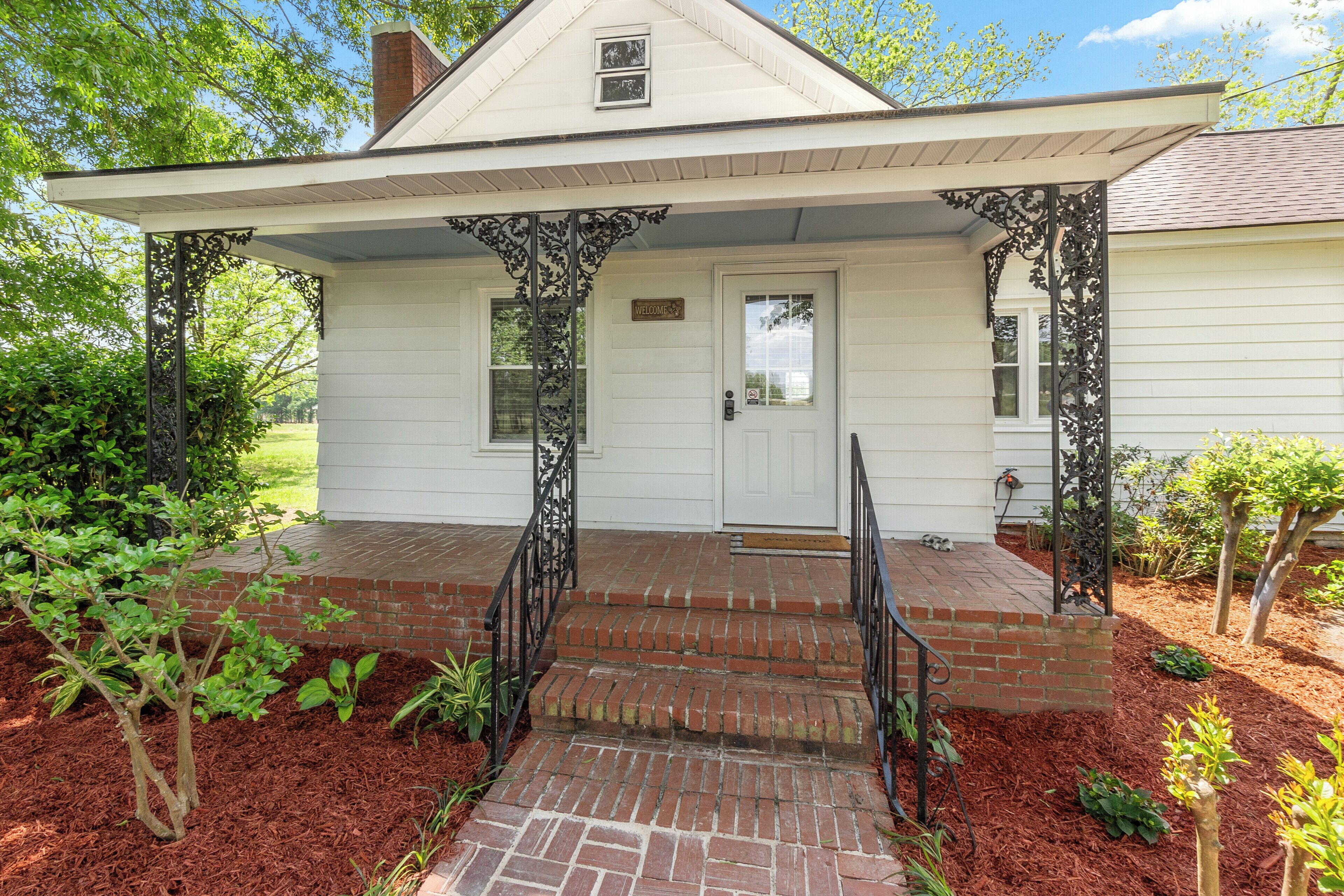 A white house with a brick porch, a front door, and a small garden area with mulch and plants.
