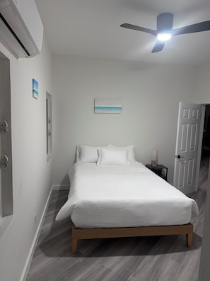 Un dormitorio con una cama, un ventilador de techo, una mesita de noche con lámpara y una puerta.
