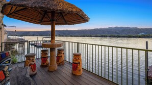 Een terras met een rieten parasol, houten krukken en uitzicht op een rivier en bergen.