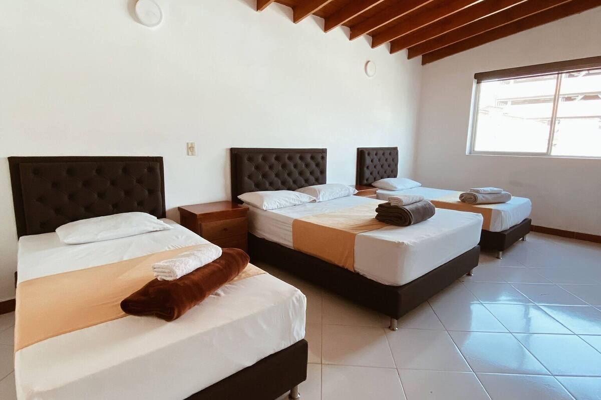 A hotel room with three beds, wooden ceiling beams, white walls, and a large window.