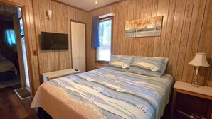 A bedroom with a large bed, wooden walls, a mounted TV, and a window with a view. - Dewitt -Cozy cabin in the woods- perfect for a small family and a dog near SML State Park (Huddleston)