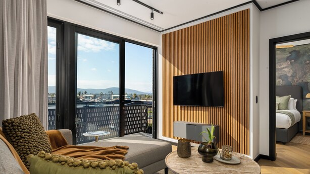 Ein modernes Hotelzimmer mit großzügigem Balkon, einem Flachbildfernseher an einer Wand aus Holzpaneelen und einer gemütlichen Sitzecke mit Sofa und rundem Couchtisch.