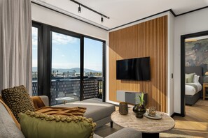 A modern hotel room with a large balcony, a flat-screen TV mounted on a wooden panel wall, and a cozy seating area with a sofa and a round coffee table. - Designer Century City Apartment - New Development (Cape Town)