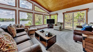 A spacious living room with a large window overlooking a lake, furnished with leather sofas, a coffee table, and a television. - Flip Flop Hideaway- Sandy beach for kids huge yard for dogs and fire pit for smores! (Huddleston)