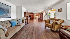 A spacious living room with a wooden floor, a large painting on the wall, and a view into the dining area. - Flip Flop Hideaway- Sandy beach for kids huge yard for dogs and fire pit for smores! (Huddleston)