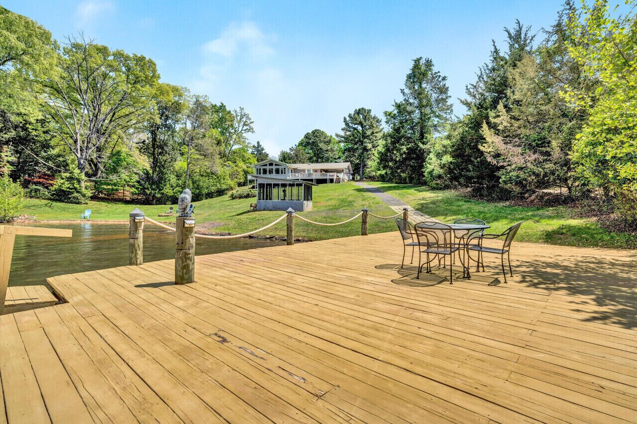 Une terrasse en bois avec une enceinte en verre, une table et des chaises, et une vue sur une maison située de l’autre côté d’une étendue d’eau.