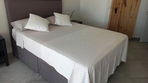 Una cama bien tendida con una colcha blanca y dos almohadas, un cabecero de madera, una mesita de noche con una lámpara y una puerta de madera al fondo.