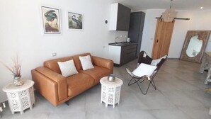 Una sala de estar moderna con un sofá marrón, dos mesas auxiliares, una silla blanca y una cocina al fondo.