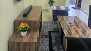 Una sala de estar con un televisor de pantalla plana montado en la pared, un banco de madera con una planta en maceta, un sofá negro y una mesa de centro.