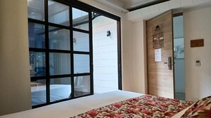 A hotel room with a large window, a bed with a floral bedspread, and a wooden door with a plaque. - POMAROSA (Medellín)