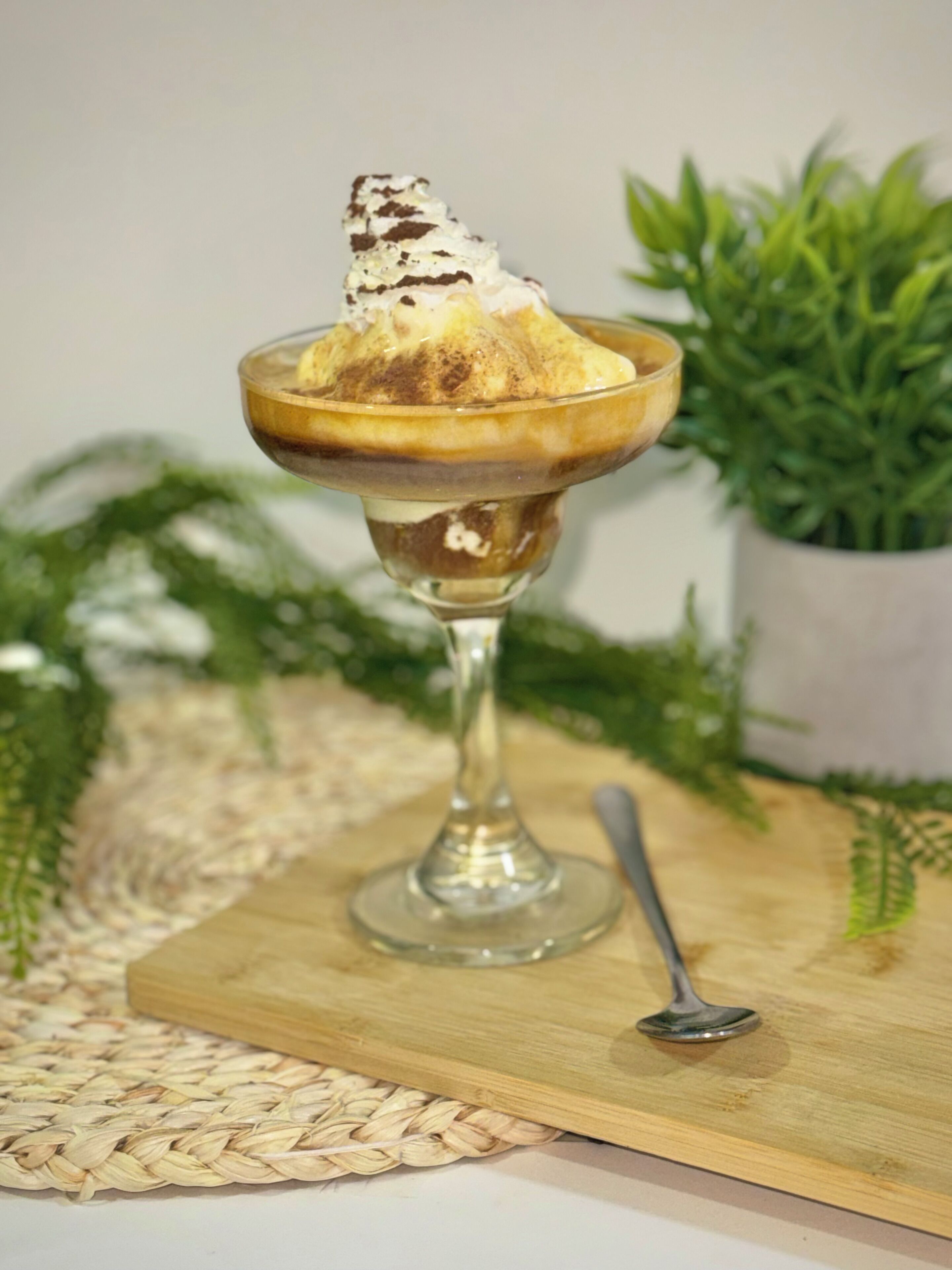A dessert in a glass bowl on a wooden board with a spoon, surrounded by greenery.