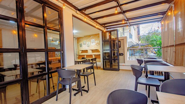 An indoor dining area with tables and chairs, large windows, and a brick wall. - Novus Laureles (Medellín)