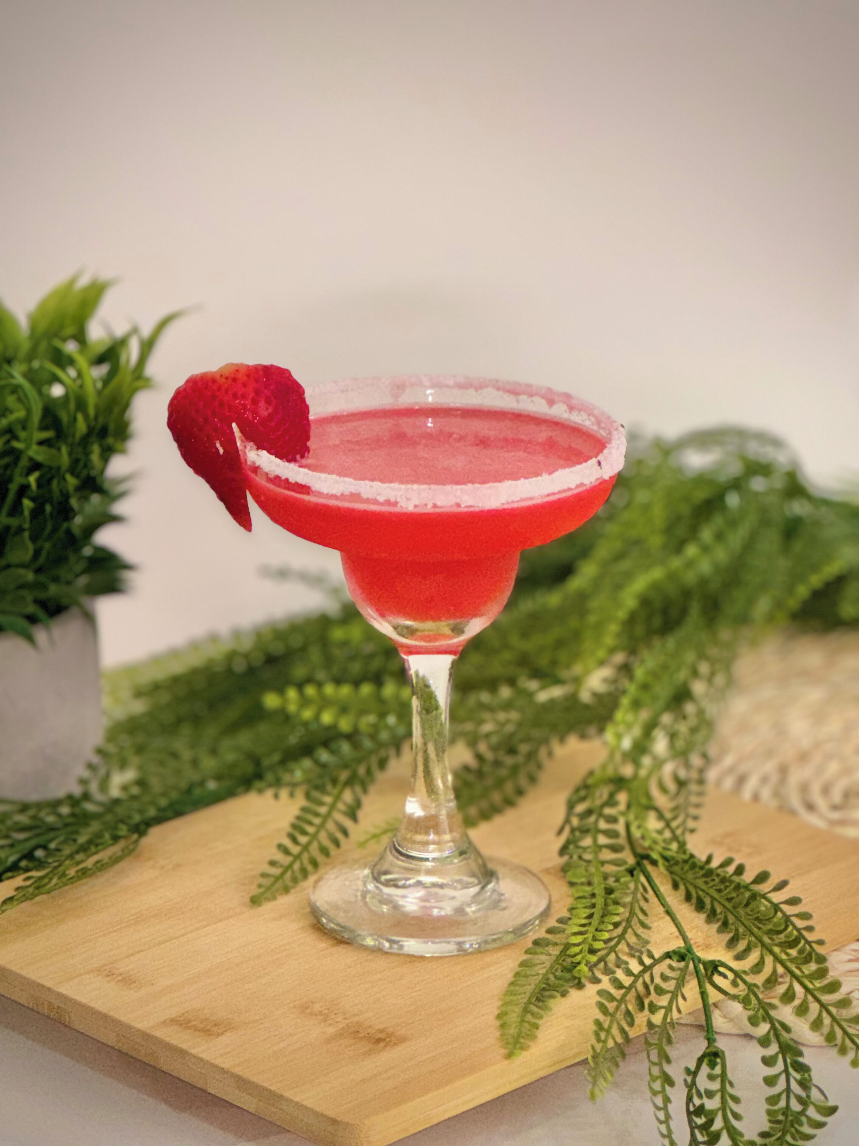 Een cocktailglas met een rode drank, versierd met een aardbei, op een houten plank met groene begroeiing.