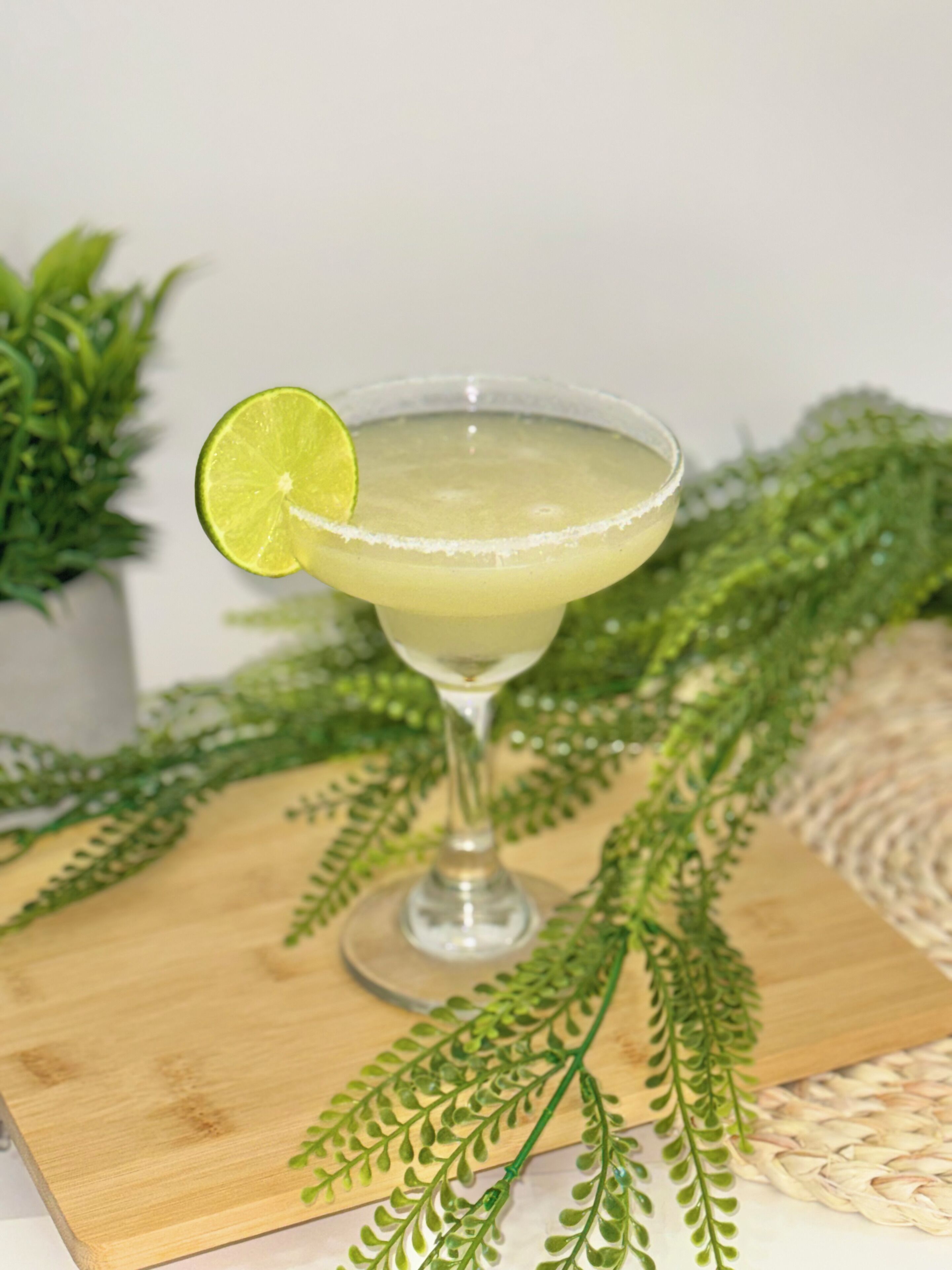 Een cocktailglas met een limoenpartje op een houten oppervlak, omringd door groen gebladerte.