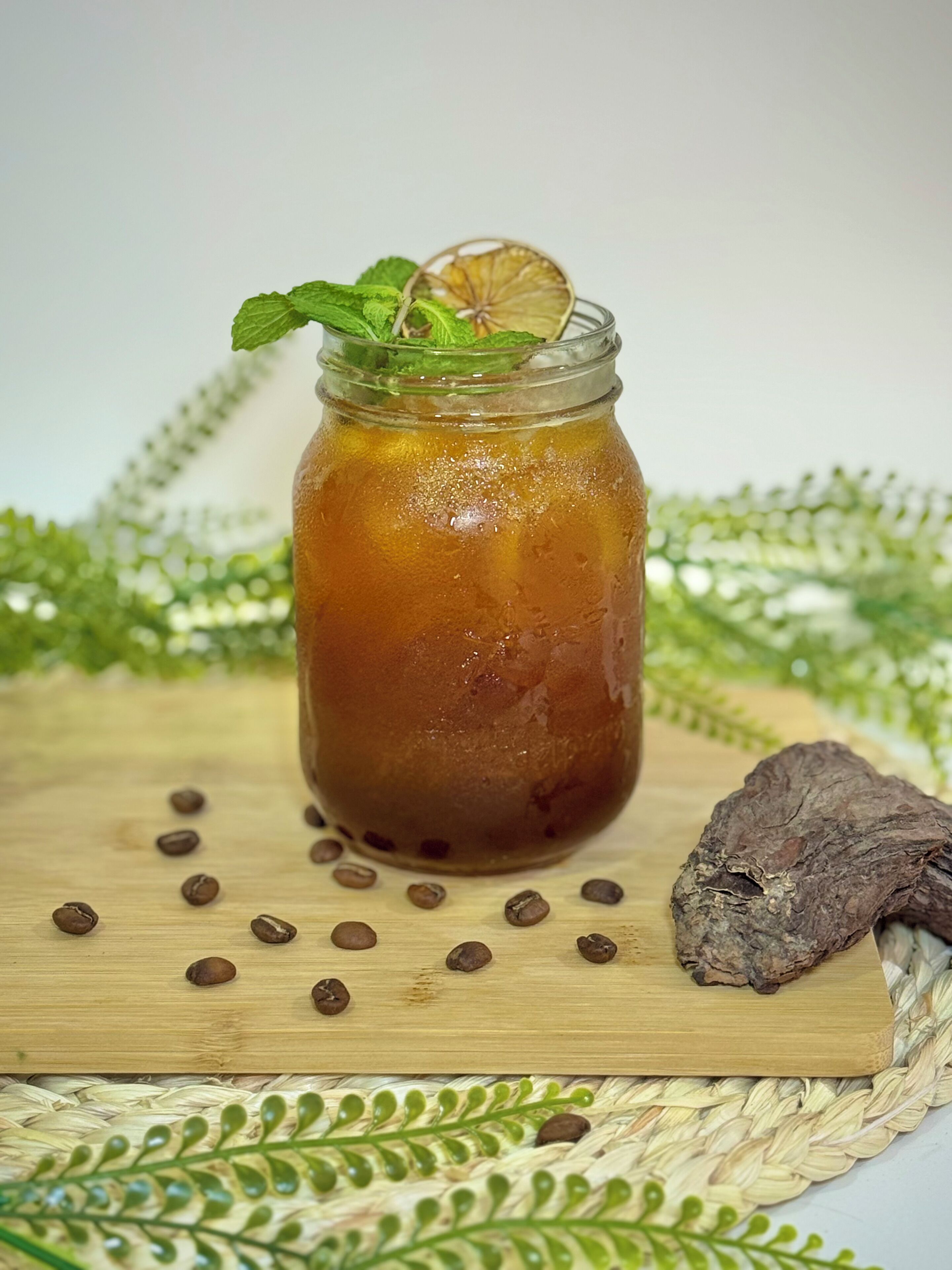 A glass jar with a brown liquid, mint leaves, and a dried orange slice on top, surrounded by coffee beans and a dried root on a wooden board.
