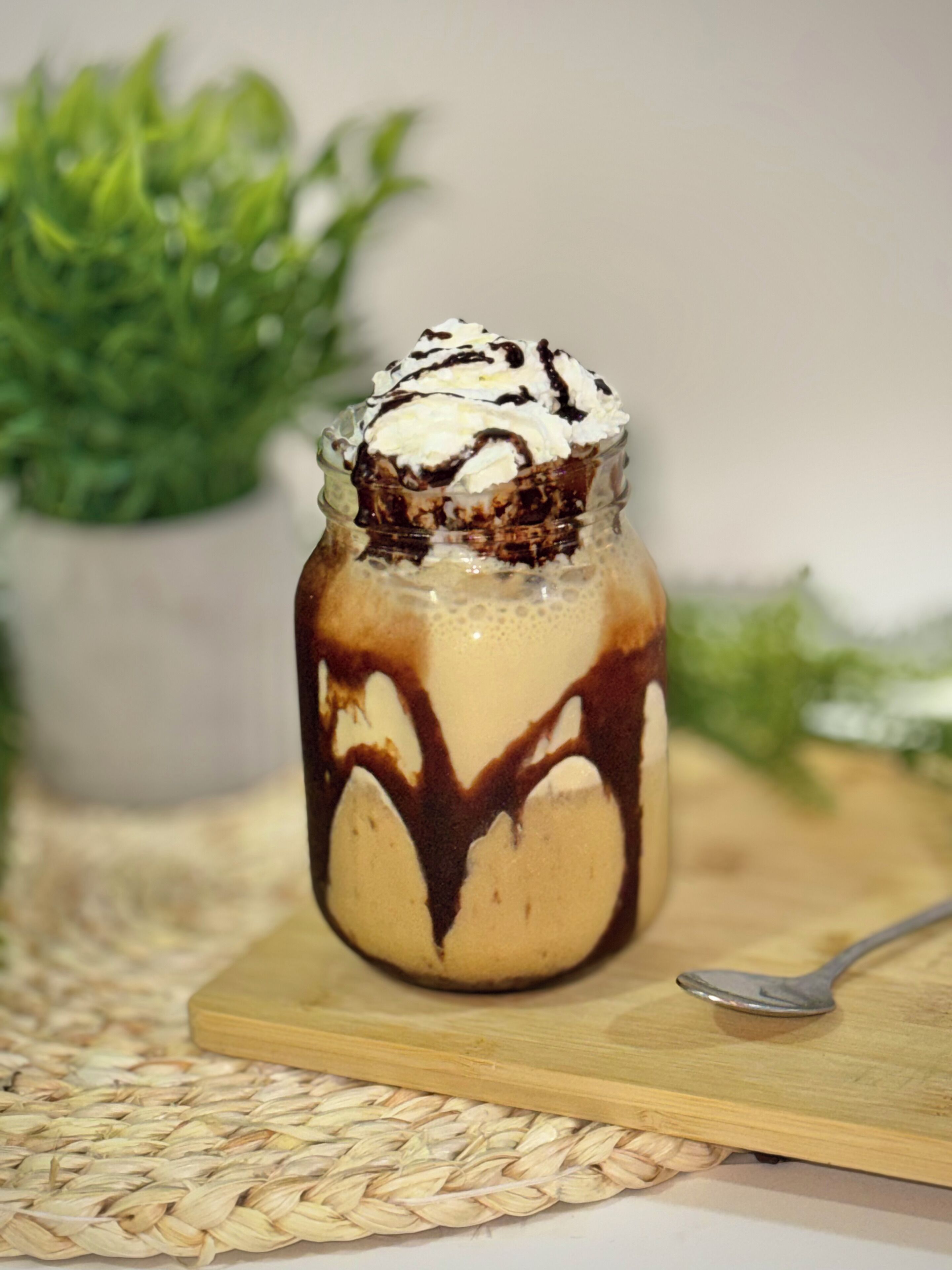 A dessert in a glass jar with chocolate sauce and whipped cream on top, placed on a wooden board with a spoon beside it.