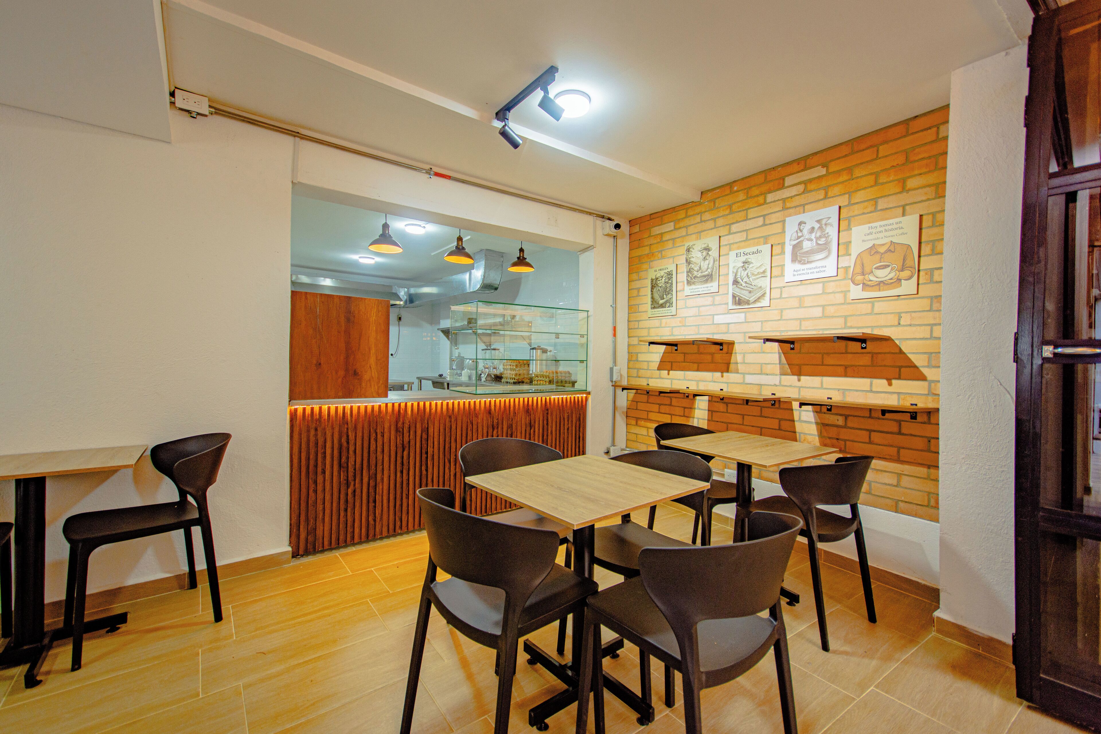 A restaurant with wooden tables and chairs, a brick wall with framed pictures, and a kitchen area in the background.