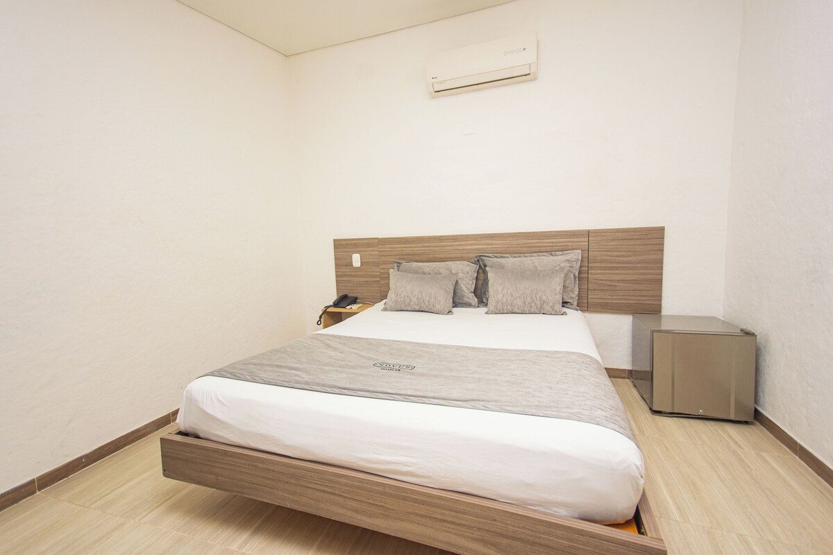 A modern hotel room with a wooden headboard, a bed with grey bedding, a bedside table, and a small bedside heater.