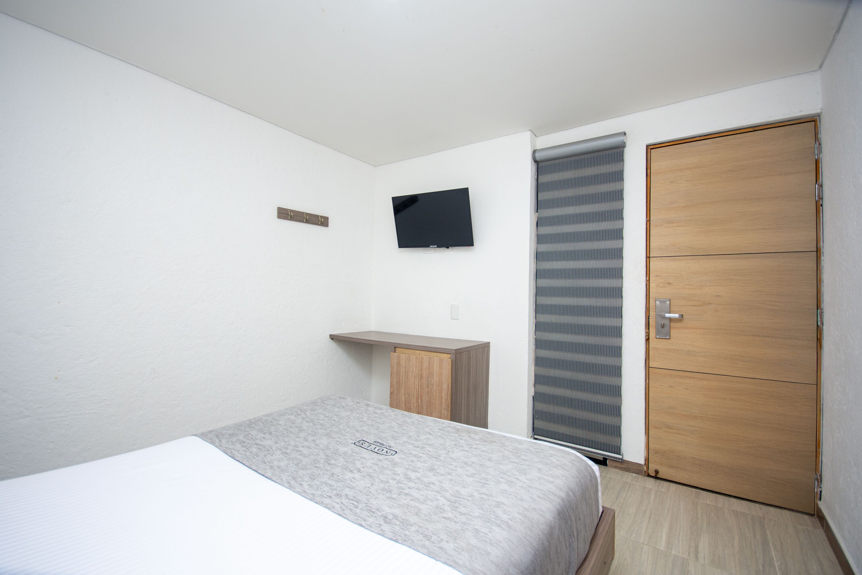 A bedroom with a bed, a TV mounted on the wall, a wooden wardrobe, and a window with blinds.