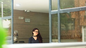 A person standing in front of a building with a check-in/out sign, a security camera, and a glass door. - Novus Laureles (Medellín)