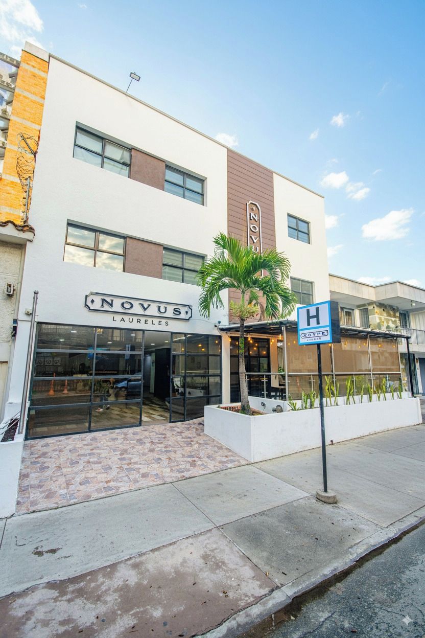 A hotel named 'Novus Laureles' with a modern facade, a covered outdoor seating area, and a 'H' sign indicating a hotel.