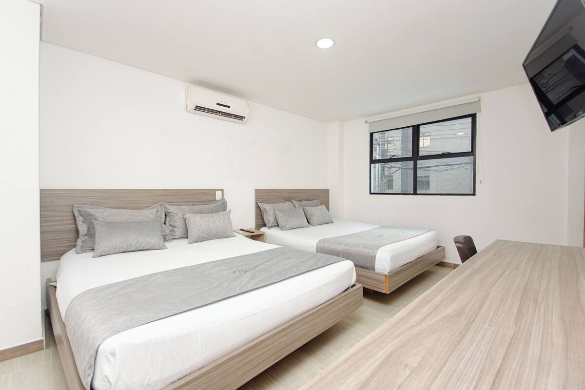A modern hotel room with two single beds, a wooden floor, a large window with a city view, and a flat-screen TV mounted on the wall.
