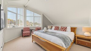 A bedroom with a sloped ceiling, a large bed with pillows and a blanket, a chair, a bedside table with a lamp, and a window with blinds. - Parkwood 5 - 2BR Split Level l Spa Bath l BBQ l Central Location l WiFi (Jindabyne)