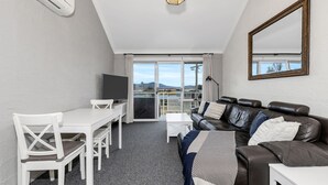A modern living room with a white table, chairs, a leather sofa, and a TV. - Parkwood 5 - 2BR Split Level l Spa Bath l BBQ l Central Location l WiFi (Jindabyne)